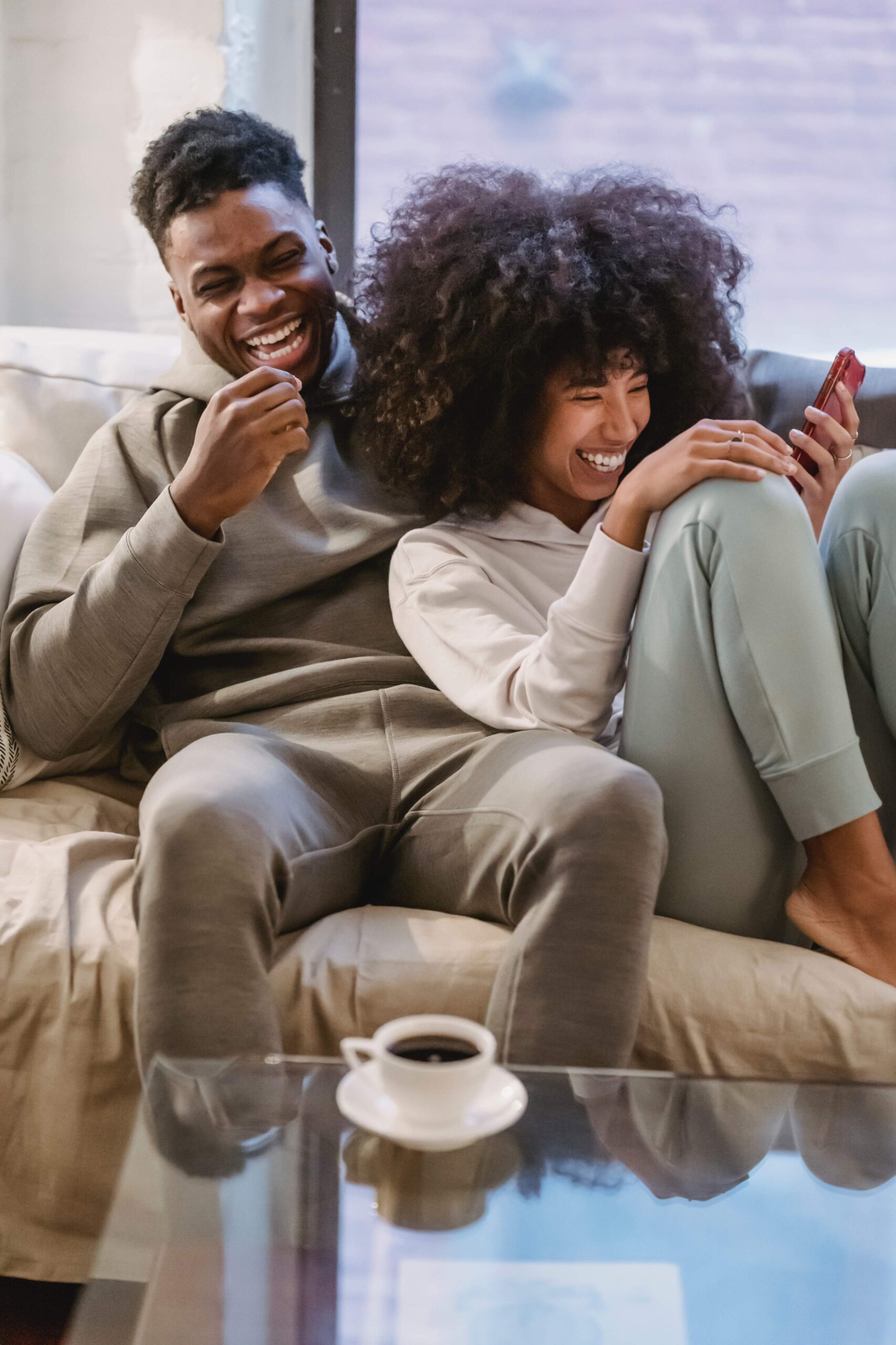 A smiling couple sits closely together on a couch, laughing and looking at a phone. The man wears a gray hoodie and sweatpants; the woman, also in loungewear, holds the phone. A cup of coffee is on the table in front of them.